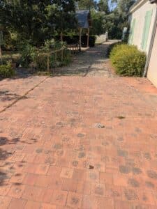 Terrasse en carreaux de terre cuite tachés menant à une allée de pavés, bordée d'un cabanon et d'une maison aux volets verts.