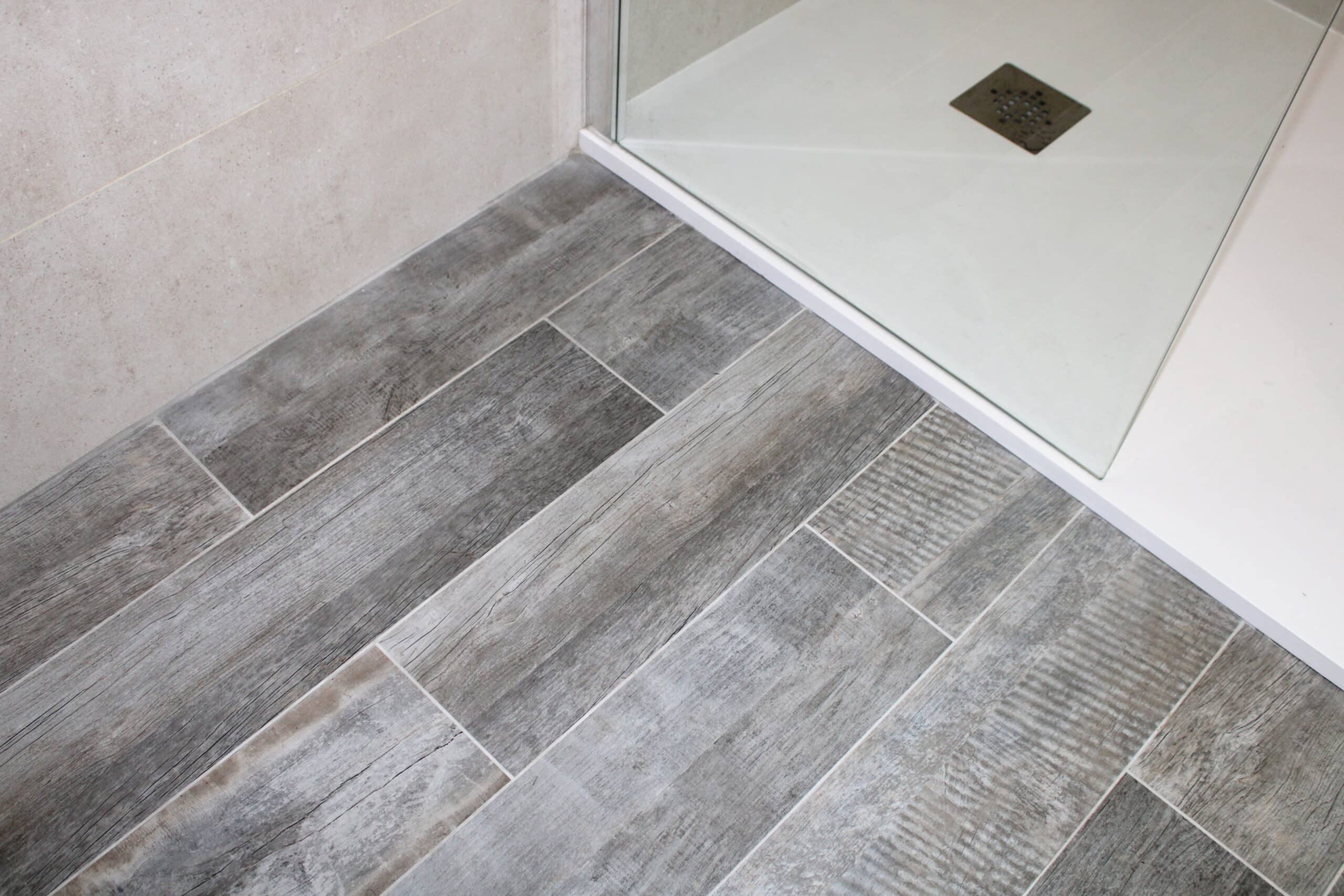 Carrelage imitation bois gris posé en diagonale devant un receveur de douche blanc avec grille d'évacuation carrée.