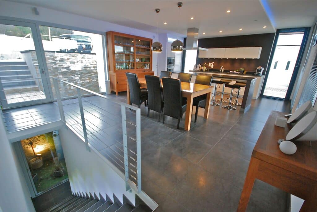 Intérieur moderne avec cuisine ouverte, salle à manger moderne (table bois, chaises noires) et escalier avec garde-corps métallique.