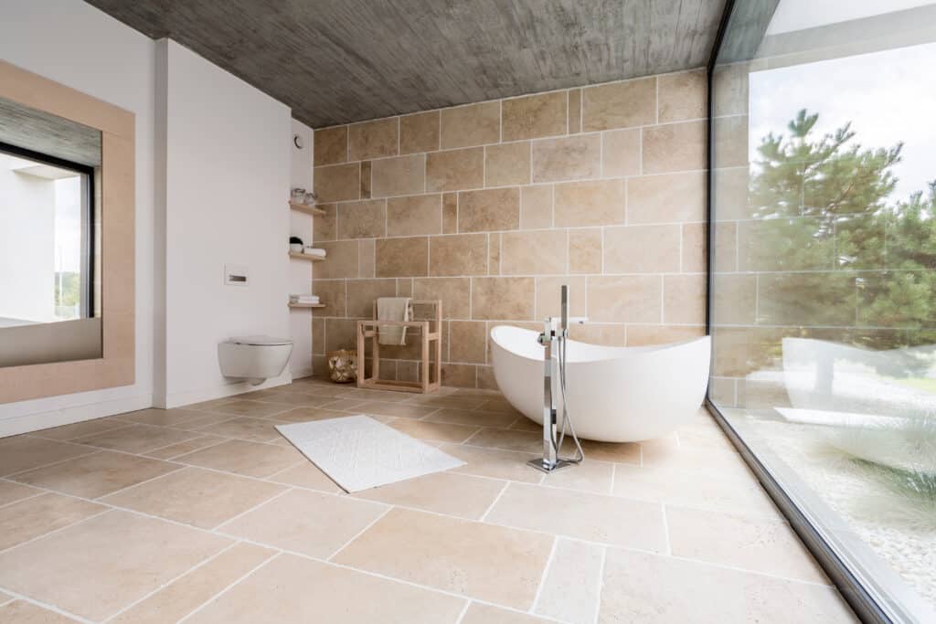 Salle de bain moderne avec baignoire îlot, carrelage en travertin, mur de pierre et grande fenêtre donnant sur la verdure.
