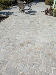 Terrasse en dalles de pierre naturelle carrées grises et beiges, avec mobilier de jardin en bois et ombres projetées au soleil.